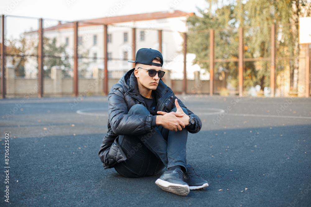 Stylish handsome young man model with sunglasses with a cap in a fashion winter down jacket with sneakers sits on the street at the stadium Stylish handsome young man model with sunglasses with a cap in a fashion winter down jacket with sneakers sits on the street at the stadium