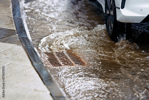Cuadro en lienzo Flood water rushing over road and into storm drain grate, car driving along flooded road during rain
