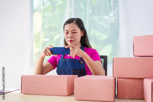 A woman in overalls sells products online, inspects parcels, takes customer orders, works at a desk, and uses pink boxes to pack items ready for shipping to customers.