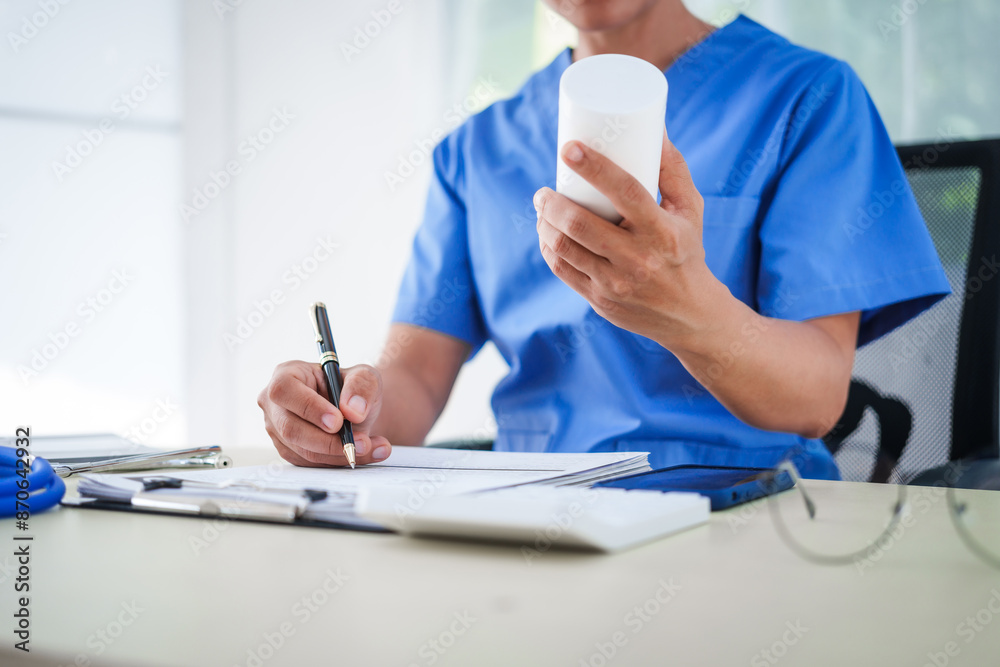 Fototapeta premium An Asian female nurse sits at a table, talking on the phone with a patient, explaining medicine during an online consultation. This faster, cheaper method helps prevent disease spread and eases