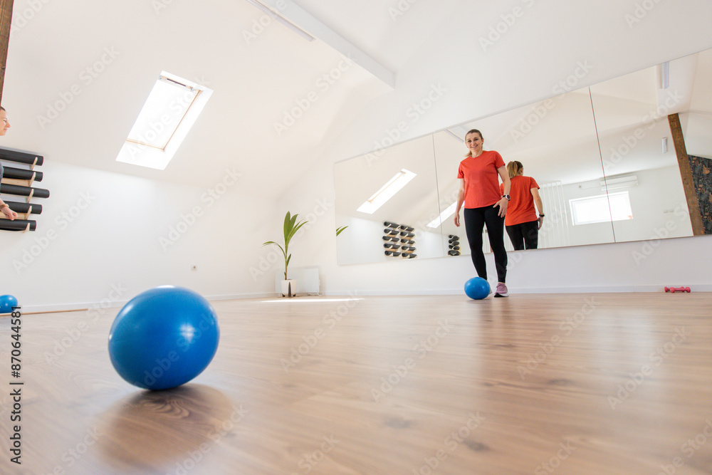 Fototapeta premium Woman In A Fitness Studio Performing An Exercise With A Blue Ball, Reflected In A Large Mirror