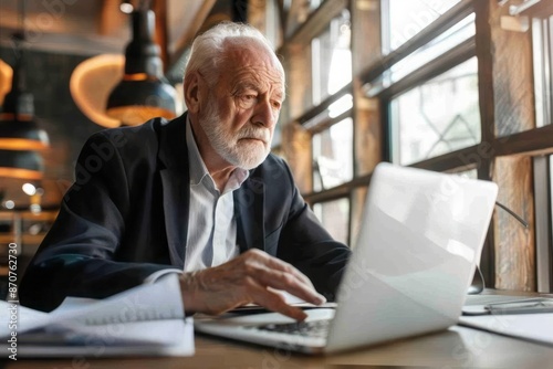 An elderly man reviewing documents next to his laptop in a sleek office setting, with a focus on his engagement in important work tasks, emphasizing his experience and expertise © DK_2020