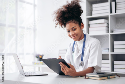 A woman wearing a white lab coat is sitting at a desk with a laptop and tablet photo
