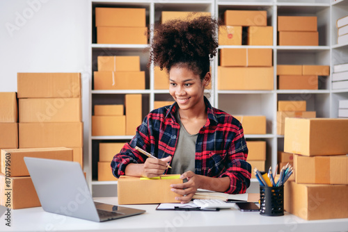 A young woman is sitting at a desk with a laptop and a stack of boxes behind her