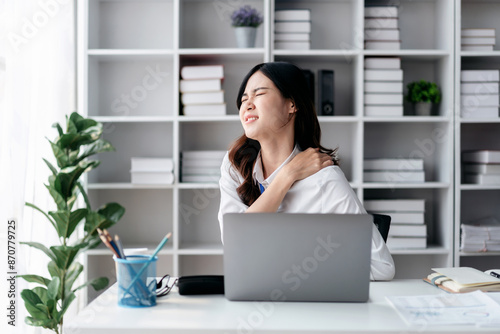 business woman is sitting at a desk with a laptop and a pain shoulder in front of her. She is injured and she is in a sad mood © Katcha