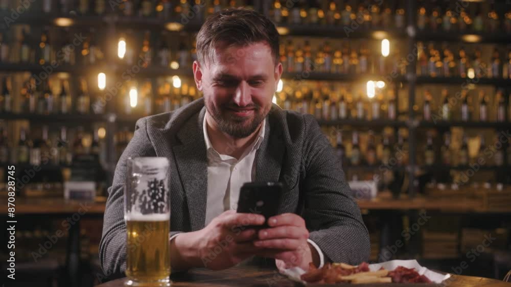 The man is at the pub, drinking beer and surfing on his phone. A handsome man is texting someone on his phone and smiling. The man is at the pub, drinking beer and surfing on his phone. A handsome man is texting someone on his phone and smiling.