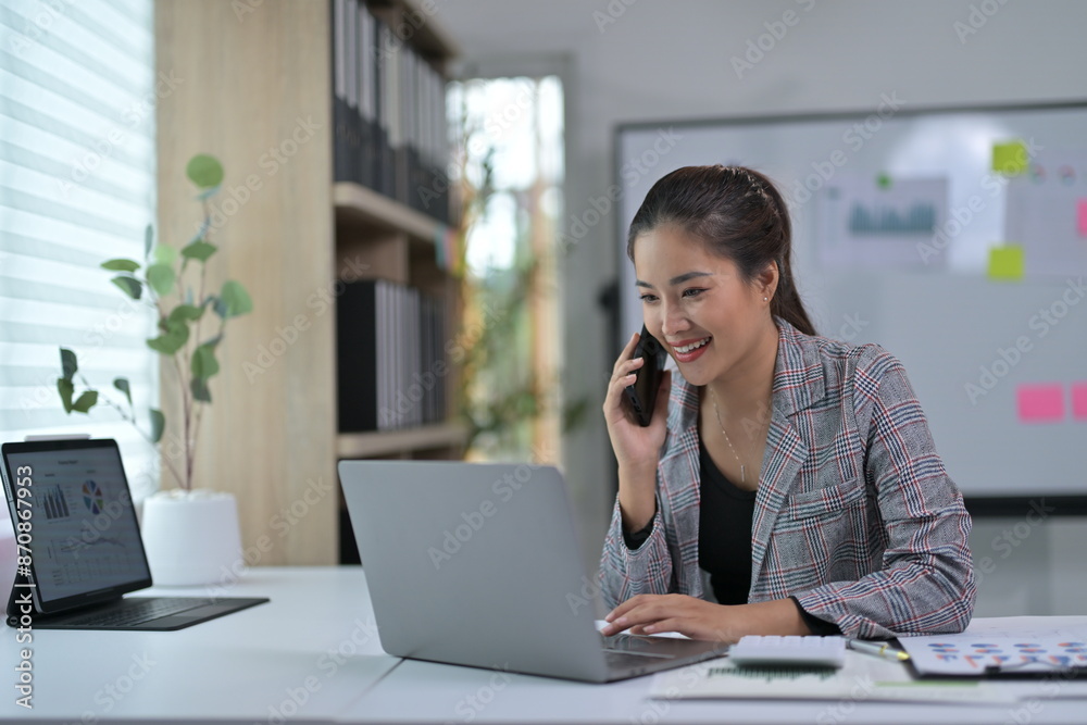 Young Professional Woman Working on Laptop and Talking on Phone in Modern Office Environment Young Professional Woman Working on Laptop and Talking on Phone in Modern Office Environment