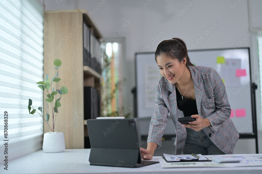 Businesswoman Working on Laptop in Modern Office with Whiteboard and Documents, Smiling and Holding Smartphone Businesswoman Working on Laptop in Modern Office with Whiteboard and Documents, Smiling and Holding Smartphone
