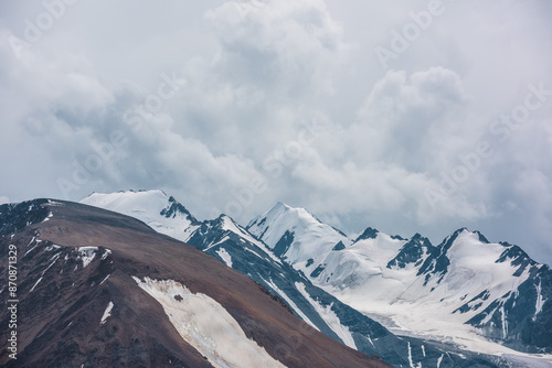 Dramatic view to large rock mountain range with snow-capped pinnacle in gray cloudy sky. Atmospheric landscape with snow cornice on snow-white pointy peak. Big rocky mountains and snowy peaked top.