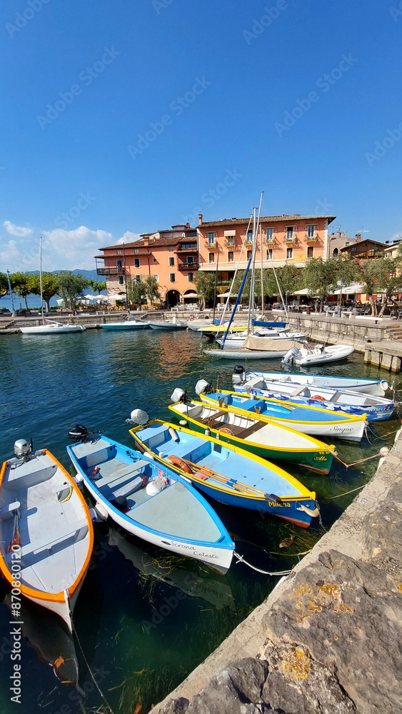 Obraz premium Colorful boats docking at a pier on a sunny day with a picturesque village in the background at lake, Garda