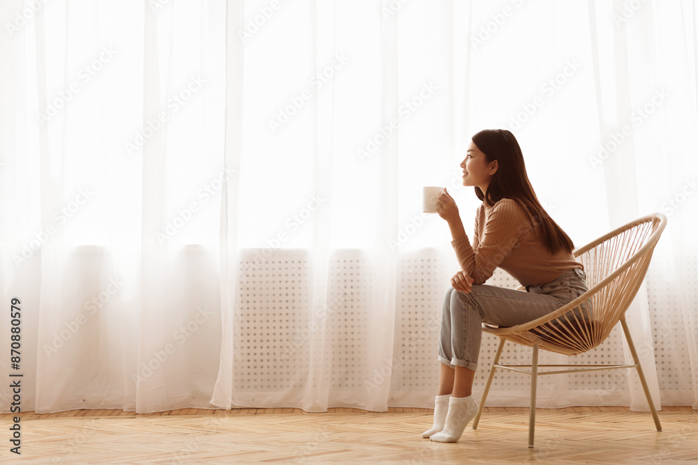 Young Girl Sitting In Modern Chair, Enjoying Coffee In Front Of Window, Side View Young Girl Sitting In Modern Chair, Enjoying Coffee In Front Of Window, Side View