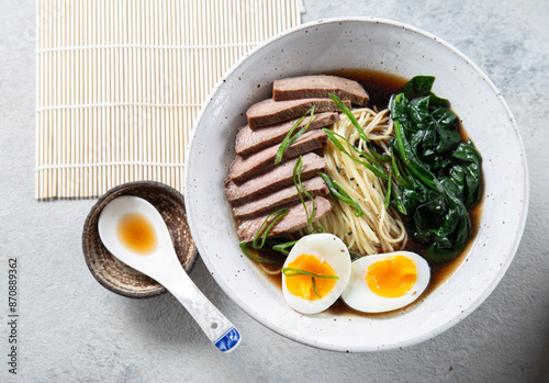 A bowl of ramen with slices of beef, soft-boiled egg halves, and green vegetables. The meal is garnished with green onions photo