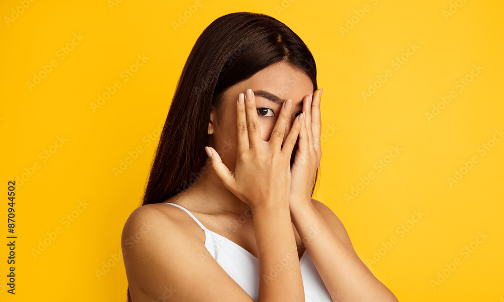 A young Asian woman with long, dark hair covers her face with her hands in a gesture of shyness or embarrassment. She is standing in front of a bright yellow background A young Asian woman with long, dark hair covers her face with her hands in a gesture of shyness or embarrassment. She is standing in front of a bright yellow background