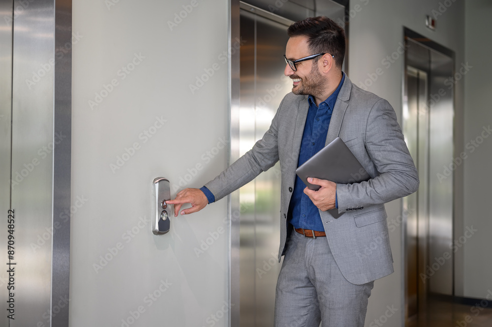 Smiling male financial advisor with laptop pushing button and waiting for elevator in modern office Smiling male financial advisor with laptop pushing button and waiting for elevator in modern office