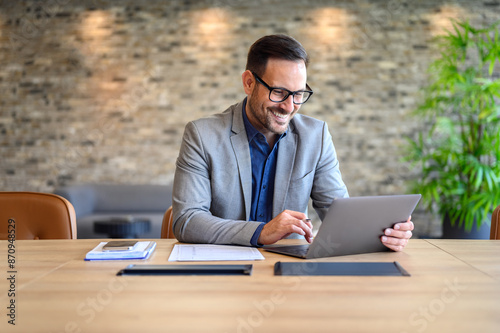 Handsome IT engineer in eyeglasses smiling and developing website over laptop on desk in modern office