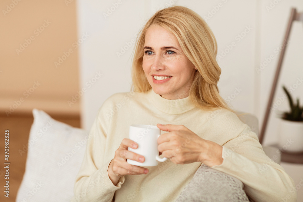 A blonde woman with a bright smile sits on a couch holding a white mug in her hands. A blonde woman with a bright smile sits on a couch holding a white mug in her hands.