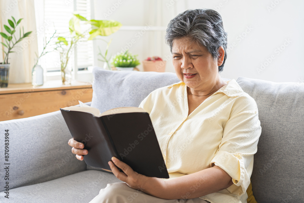 Presbyopia, Hyperopia mature, aged asian woman having vision problem trying to stare read text on a book on sofa at home, eye disease of elderly. Poor eyesight farsightedness, health care concept Presbyopia, Hyperopia mature, aged asian woman having vision problem trying to stare read text on a book on sofa at home, eye disease of elderly. Poor eyesight farsightedness, health care concept
