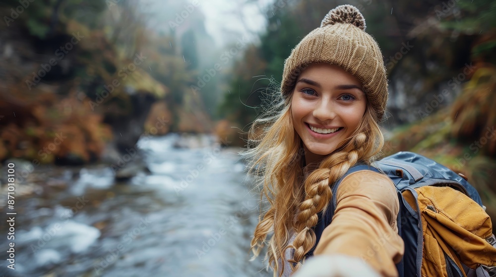 Smiling young female hiker wearing a brown knitted hat and carrying a backpack, taking a selfie while enjoying a scenic hike along a river surrounded by forested hills.