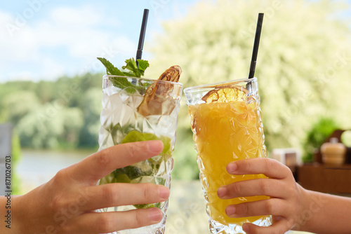 Closeup of two hands clinking cocktails against vibrant beautiful summer nature unrecognizable people drinking non alcoholic mojito and orange juice photo