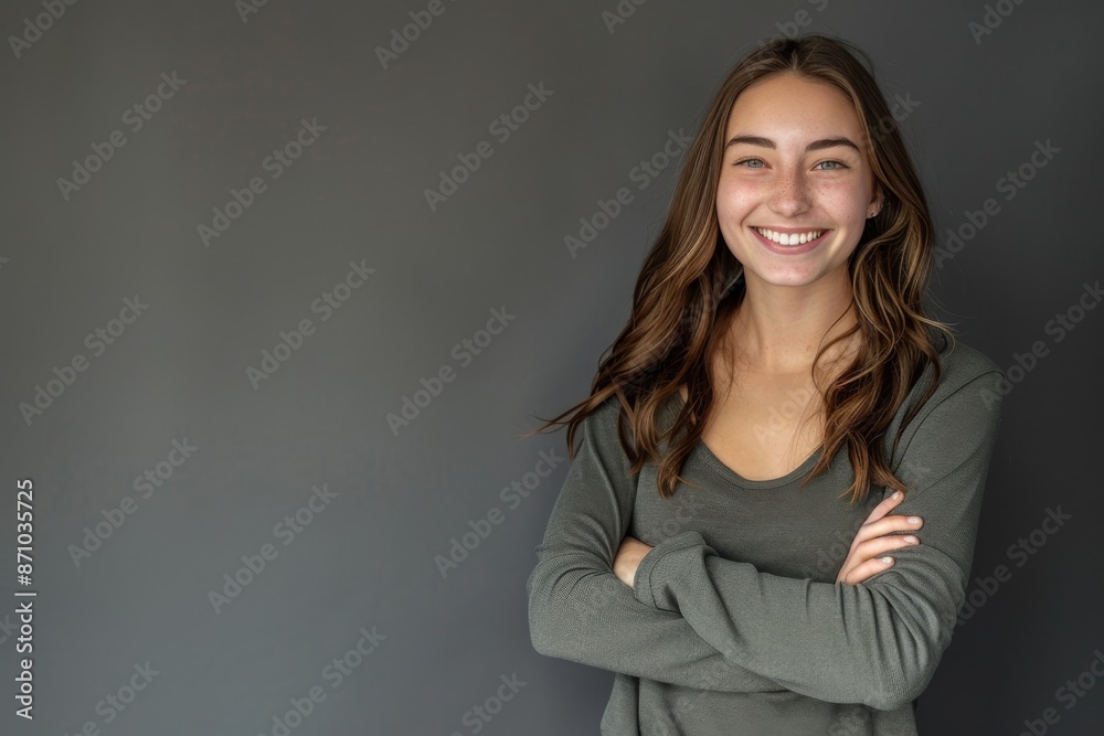 Obraz premium A portrait of a smiling young woman with long brown hair, wearing a grey sweater, posing with her arms crossed against a plain grey background