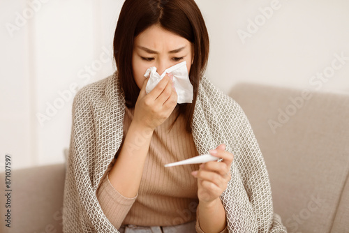 фотографія A woman is sitting on a couch at home, wrapped in a blanket and holding a thermometer