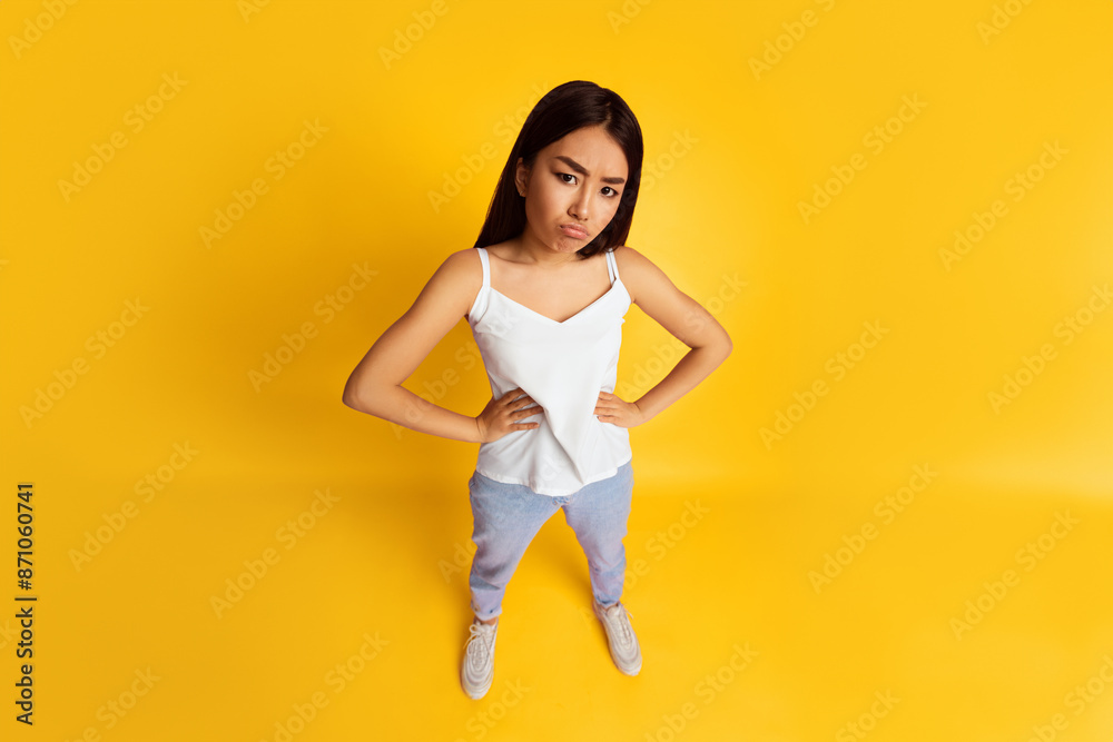 Asian young woman with dark hair standing against a bright yellow background. She is wearing a white tank top and light blue jeans. She is looking at the camera with a serious expression, top view Asian young woman with dark hair standing against a bright yellow background. She is wearing a white tank top and light blue jeans. She is looking at the camera with a serious expression, top view