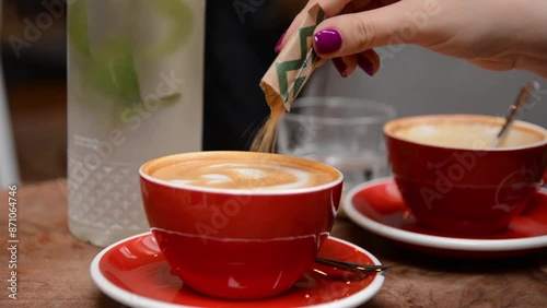 Close-up of female hand pouring brown sugar into espresso coffee in a red cup from a small packet in a cafe, with blurred background