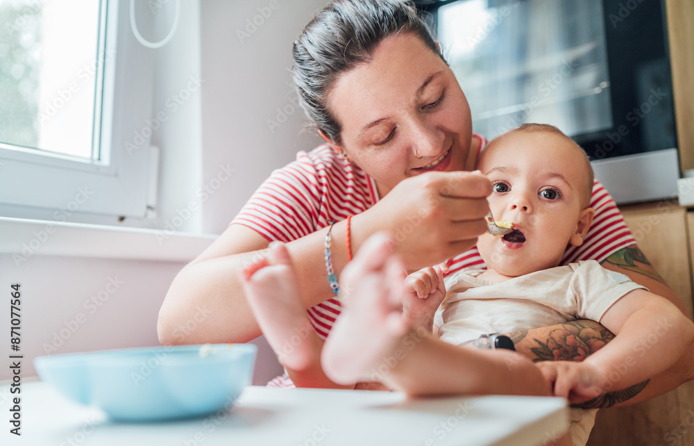 Mother lovingly feeds her baby in cozy kitchen showcasing moment of tender care and bonding. Baby, with feet playfully extended, looks content and curious, adding touch of warmth and intimacy to scene Mother lovingly feeds her baby in cozy kitchen showcasing moment of tender care and bonding. Baby, with feet playfully extended, looks content and curious, adding touch of warmth and intimacy to scene