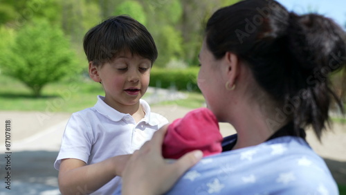 Boy looking at mother holding baby in sling, showing sibling curiosity and family bonding in a casual outdoor scene filled with natural light and greenery © Marco