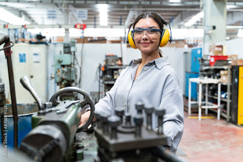 A young woman wearing safety glasses and ear protection is operating a lathe in a metalworking shop. looking at the camera and smiling. A young woman wearing safety glasses and ear protection is operating a lathe in a metalworking shop. looking at the camera and smiling.