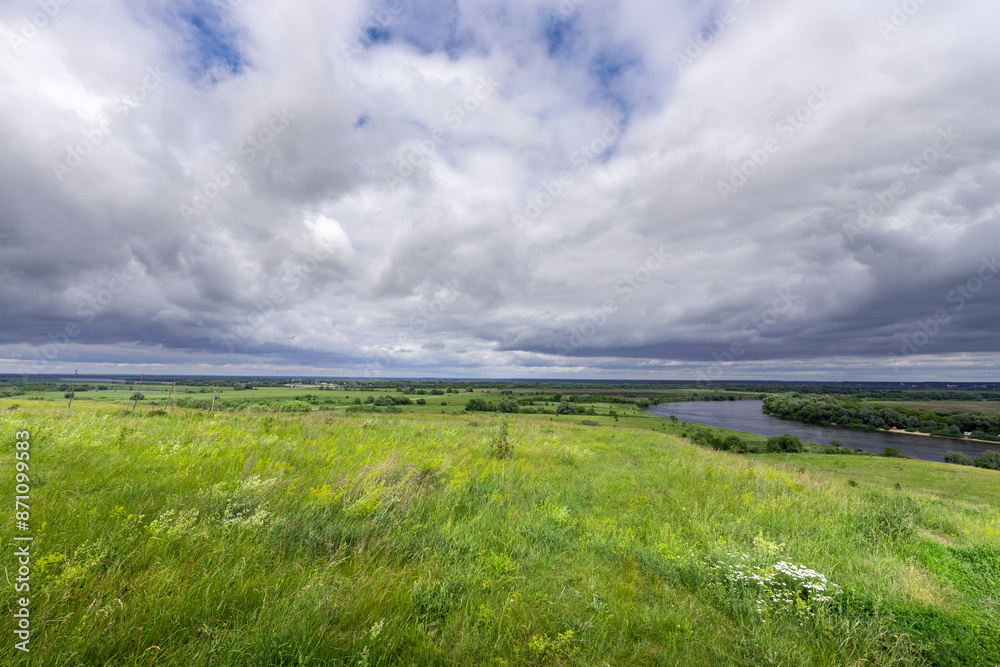 Obraz premium A panoramic view of a river valley with lush green meadows and a winding river in the distance, all under a dramatic sky filled with puffy, gray clouds.
