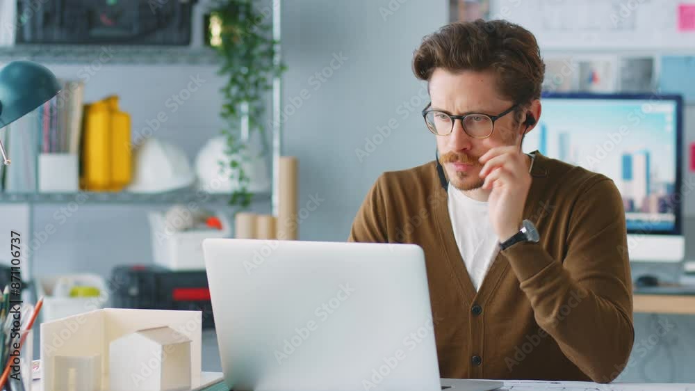 Camera tracks across frame as casually dressed architect working at laptop on desk in office listens to music on wireless earphones - shot in slow motion Camera tracks across frame as casually dressed architect working at laptop on desk in office listens to music on wireless earphones - shot in slow motion