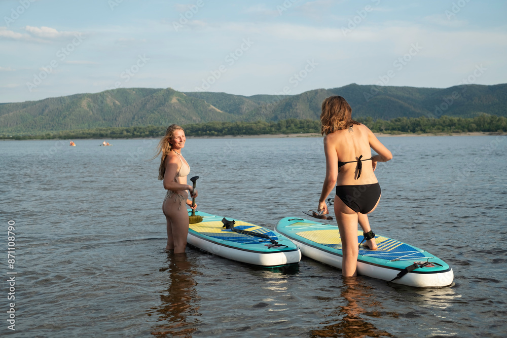 Two Women Enjoy Paddleboarding in Scenic Lake During Sunny Summer Two Women Enjoy Paddleboarding in Scenic Lake During Sunny Summer