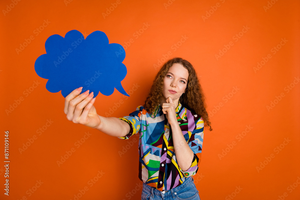 Photo of minded cute girl wear stylish print clothes hold paper card empty space isolated on orange color background Photo of minded cute girl wear stylish print clothes hold paper card empty space isolated on orange color background
