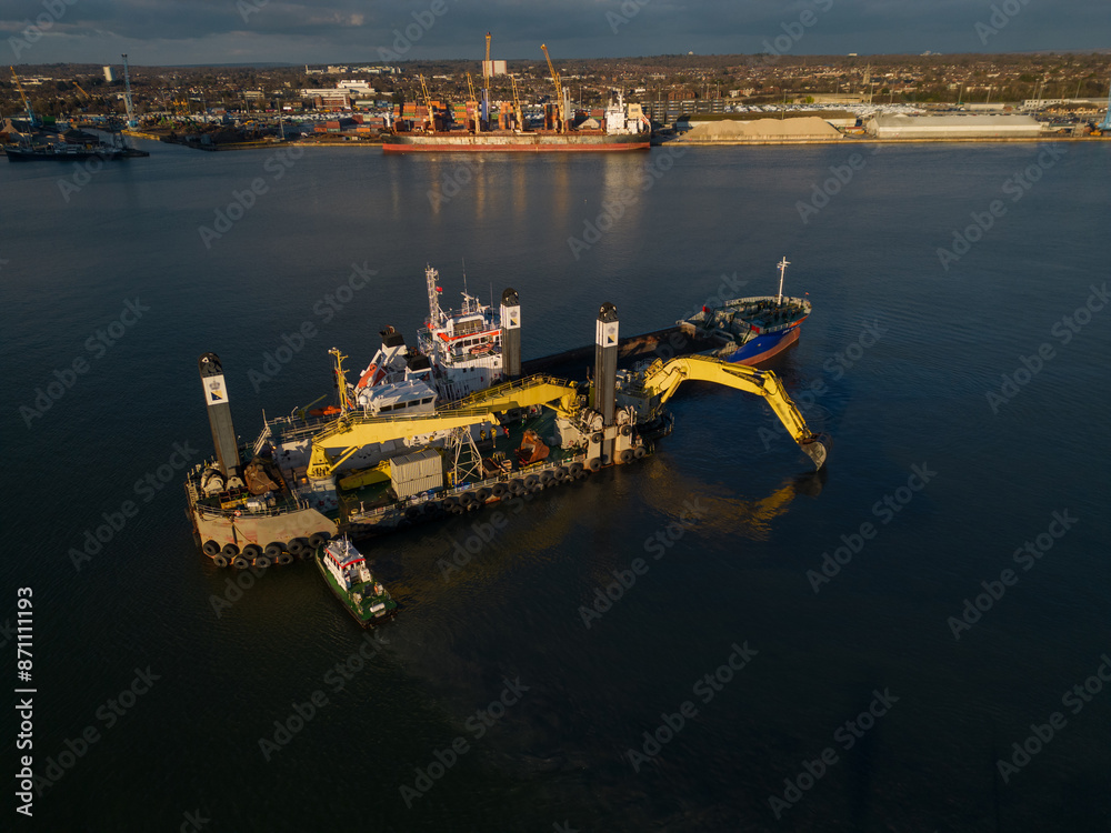 Harbour maintenance with backhoe dredger in Southampton Docks, UK. Hydraulically driven excavator on pontoon dredging. Aerial view at golden hour. Cargo ship at distance.