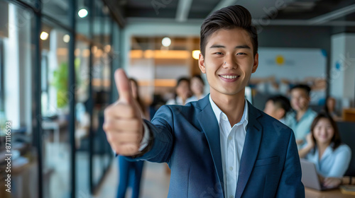 handsome Asian businessman is giving a thumbs up in the office, smiling at the camera with a group of people behind him