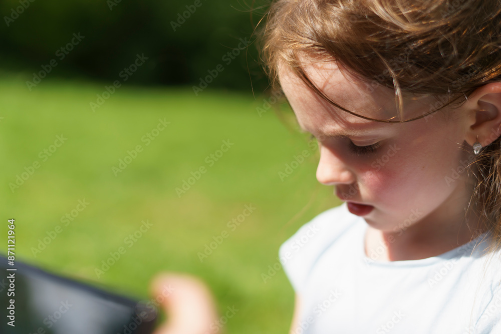 A sad little cute preschooler girl in a green clearing in the forest looks down when she is photographed on a phone. Child loneliness concept A sad little cute preschooler girl in a green clearing in the forest looks down when she is photographed on a phone. Child loneliness concept