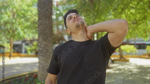 Hispanic man with a beard in a black shirt feeling neck pain in a sunny park surrounded by greenery photo