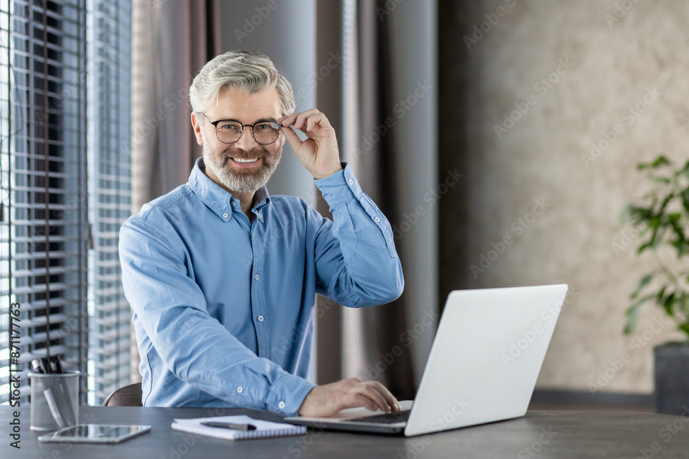 A confident middle-aged businessman wearing glasses and a blue shirt, smiling while working on his laptop in a modern office setting. The image captures professionalism, positivity, and productivity. A confident middle-aged businessman wearing glasses and a blue shirt, smiling while working on his laptop in a modern office setting. The image captures professionalism, positivity, and productivity.