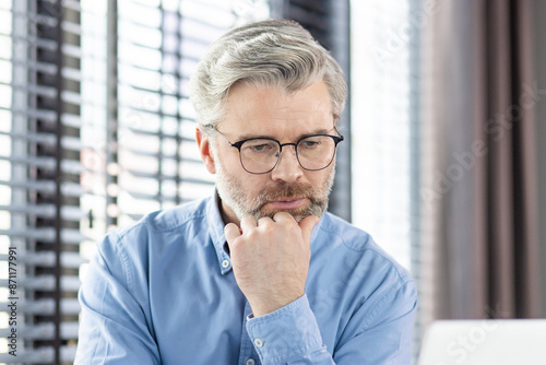 A mature businessman in glasses and a blue shirt is thinking deeply while working on a laptop in a modern office. The image conveys concentration, thoughtfulness, and dedication.