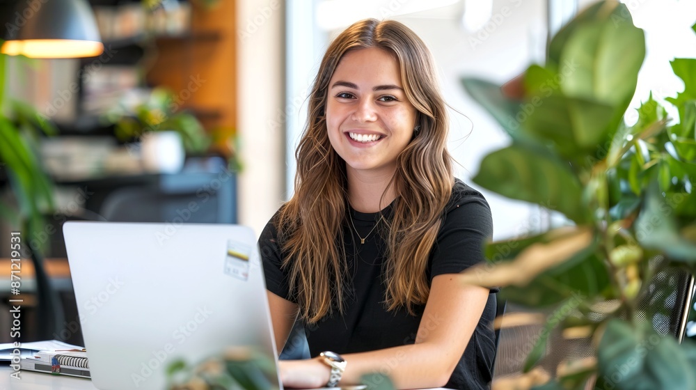 Smiling woman using laptop in modern office setting. Bright workspace with green plants and natural light. Engaging work atmosphere for increased productivity and creativity