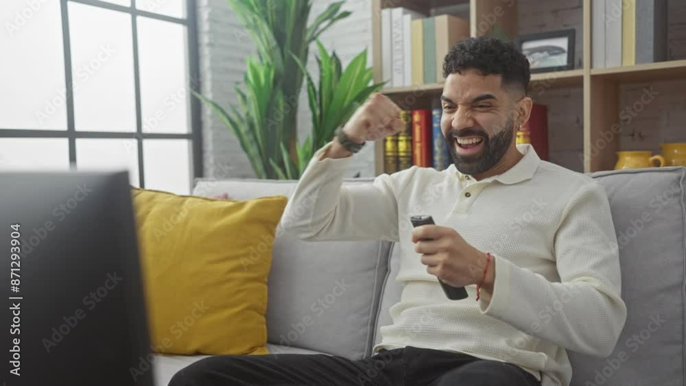 A cheerful young hispanic man with a beard celebrates while holding a remote control, seated on a sofa in a cozy living room. A cheerful young hispanic man with a beard celebrates while holding a remote control, seated on a sofa in a cozy living room.