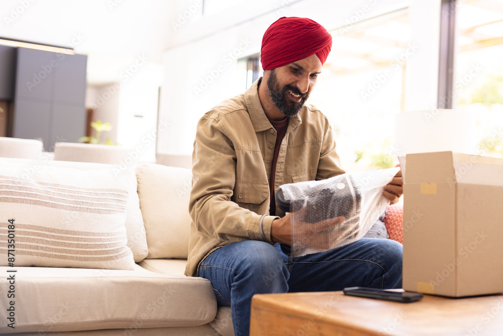 Unboxing package, man in turban sitting on sofa, smiling at new item Unboxing package, man in turban sitting on sofa, smiling at new item