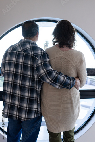 Senior couple standing together, looking out round window, hugging each other