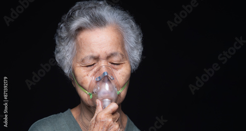 An elderly Asian woman using an oxygen mask against a black background.