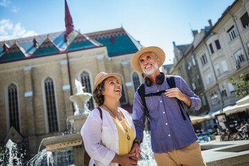 Elderly married couple exploring new city. They are wearing hats and backpacks.