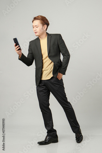 full length shoot of pensive asian business man wearing suit holding mobile phone on isolated background