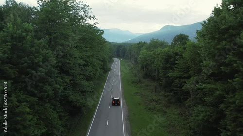 A black SUV drives along the road through a forest belt, mountains are visible in the distance