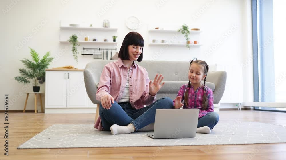 Mother and daughter sitting cross-legged on floor having video call on laptop. Smiling and waving at screen sharing joyful moment sharing fun and bonding time. Bright living room environment. Mother and daughter sitting cross-legged on floor having video call on laptop. Smiling and waving at screen sharing joyful moment sharing fun and bonding time. Bright living room environment.