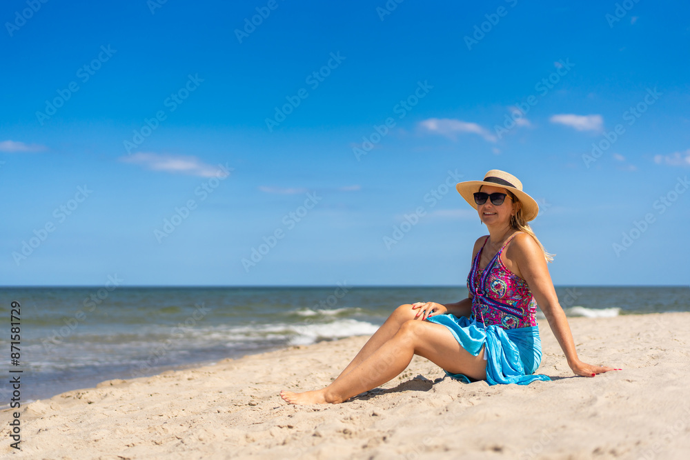 Summer holidays on the Baltic Sea. Beautiful young woman in a colorful swimsuit and a blue pareo and hat sitting on the ground on a sandy beach by the sea, looking dreamily at the horizon. Side view Summer holidays on the Baltic Sea. Beautiful young woman in a colorful swimsuit and a blue pareo and hat sitting on the ground on a sandy beach by the sea, looking dreamily at the horizon. Side view